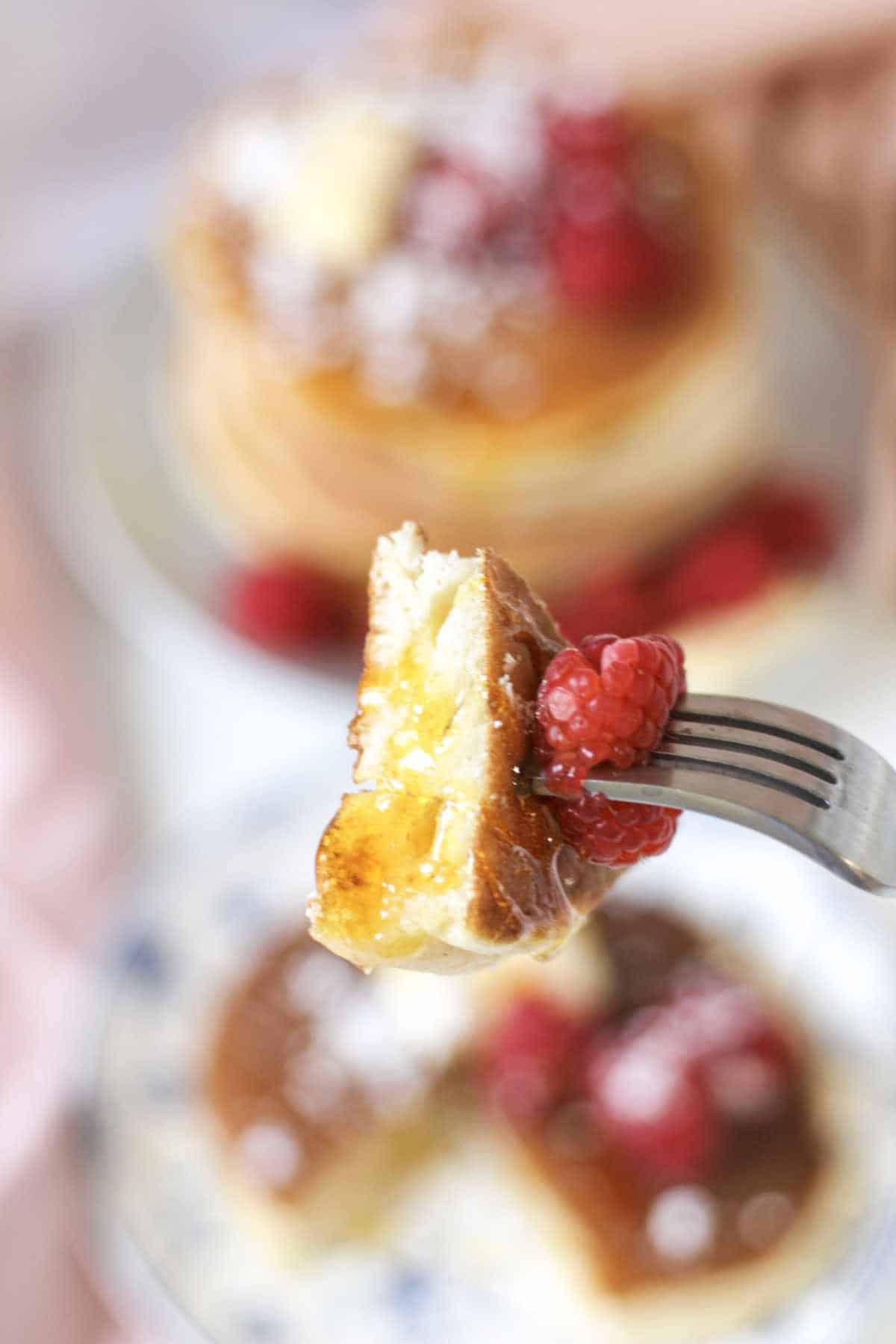 A close-up of a fork holding a bite of fluffy, old fashioned buttermilk pancake topped with powdered sugar and fresh raspberries, with a blurred stack of pancakes and berries in the background.