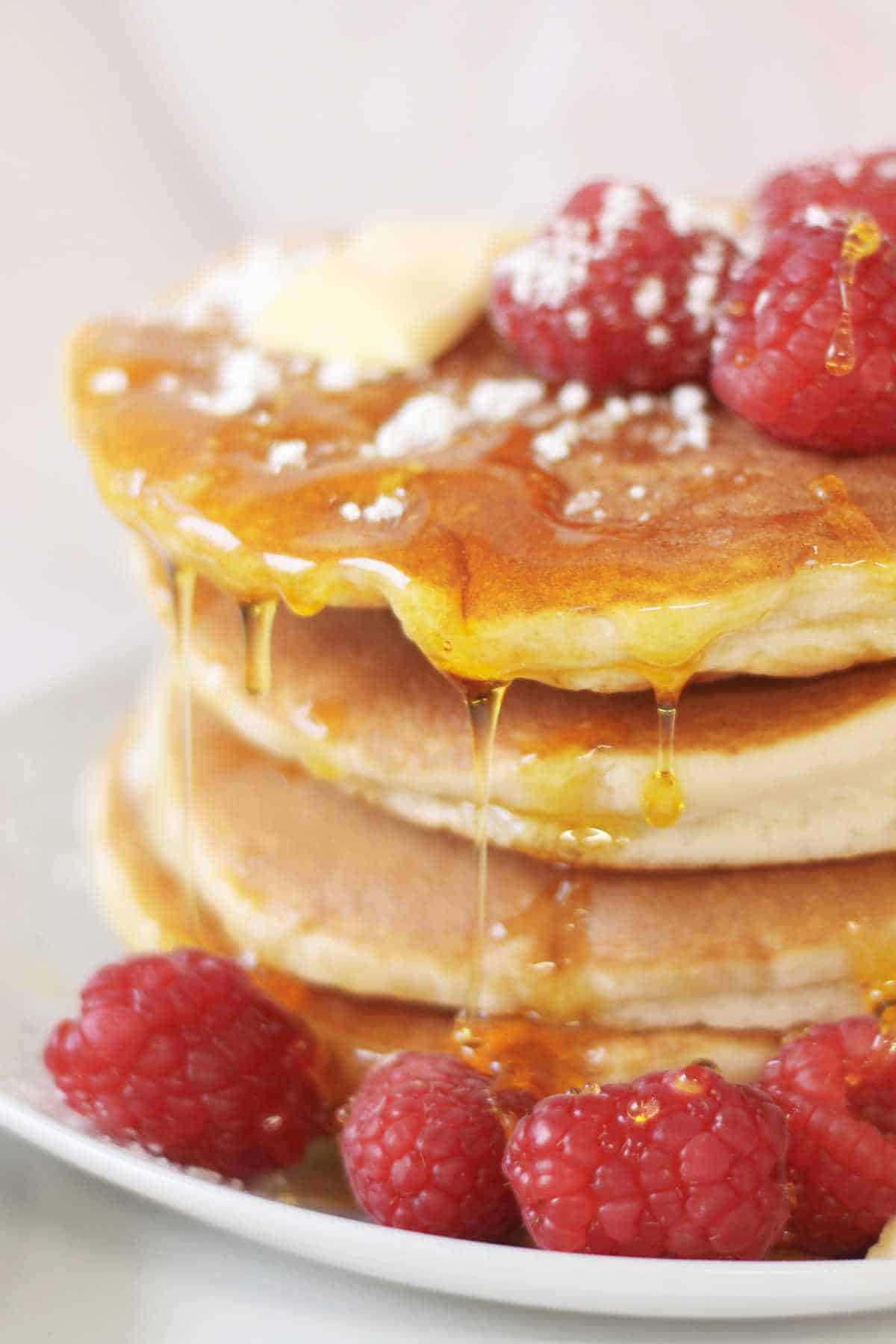 A stack of old fashioned buttermilk pancakes topped with raspberries, a pat of butter, and powdered sugar, with syrup dripping down the sides and more raspberries scattered on the plate.