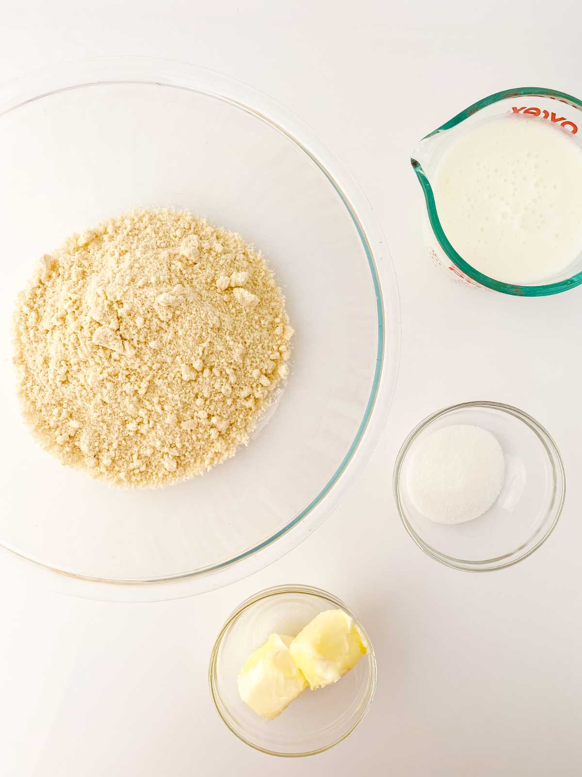 A glass bowl with flour mixture, a measuring cup of milk, a small bowl of sugar, and butter—classic ingredients arranged on a white surface for making copycat Red Lobster biscuits.