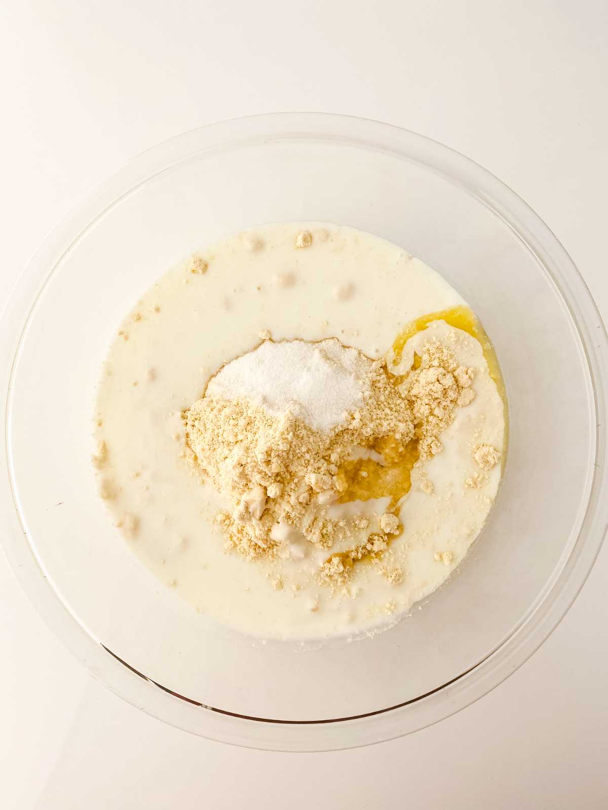 A clear glass bowl with flour, sugar, milk, and melted butter for making cheddar cheese drop biscuits sits un-mixed on a white surface.