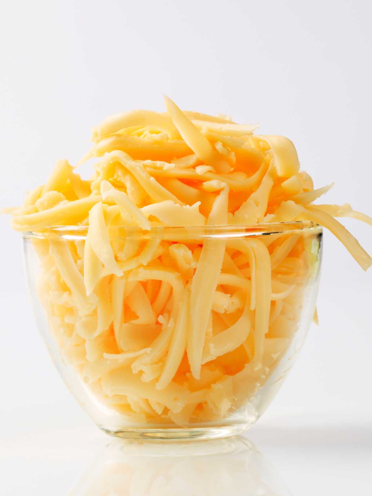 A clear glass bowl filled with a large amount of yellow shredded cheese sits on a white surface, perfect for making cheddar bay biscuits, against a plain white background.