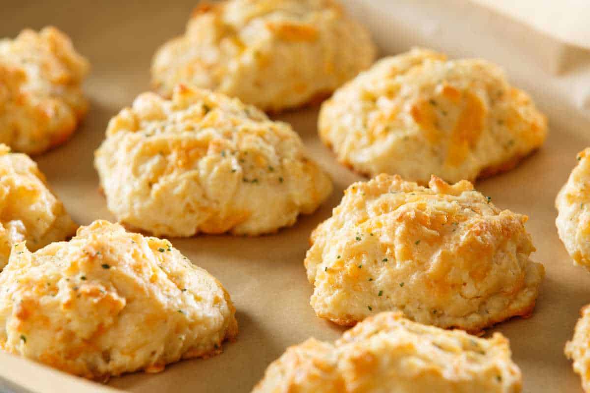 Nine golden-brown cheddar cheese drop biscuits with flecks of herbs rest on parchment paper, freshly baked and lightly textured, with melted cheese visible in the dough—just like copycat Red Lobster biscuits.