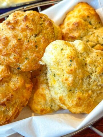 A basket lined with white paper holds several golden-brown, fluffy cheddar bay biscuits sprinkled with herbs.