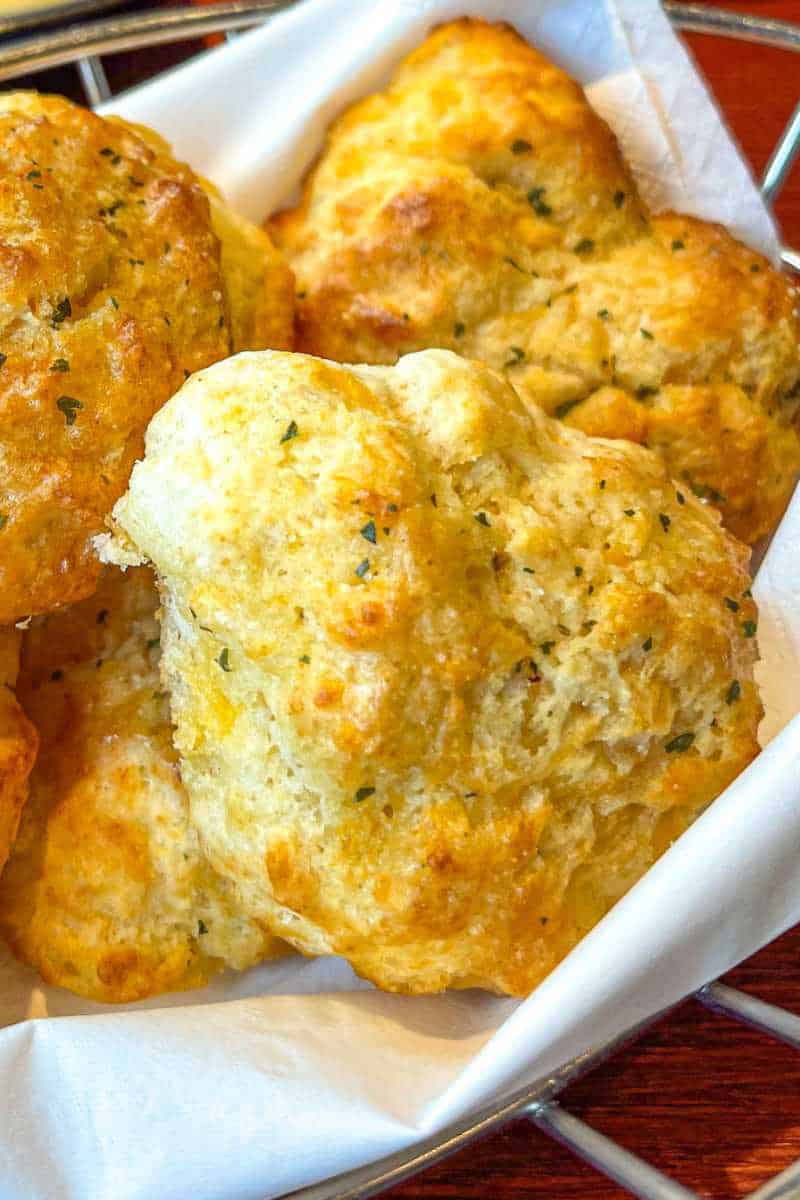 A close-up of golden, flaky cheddar bay biscuits with flecks of herbs, served in a basket lined with white parchment paper.