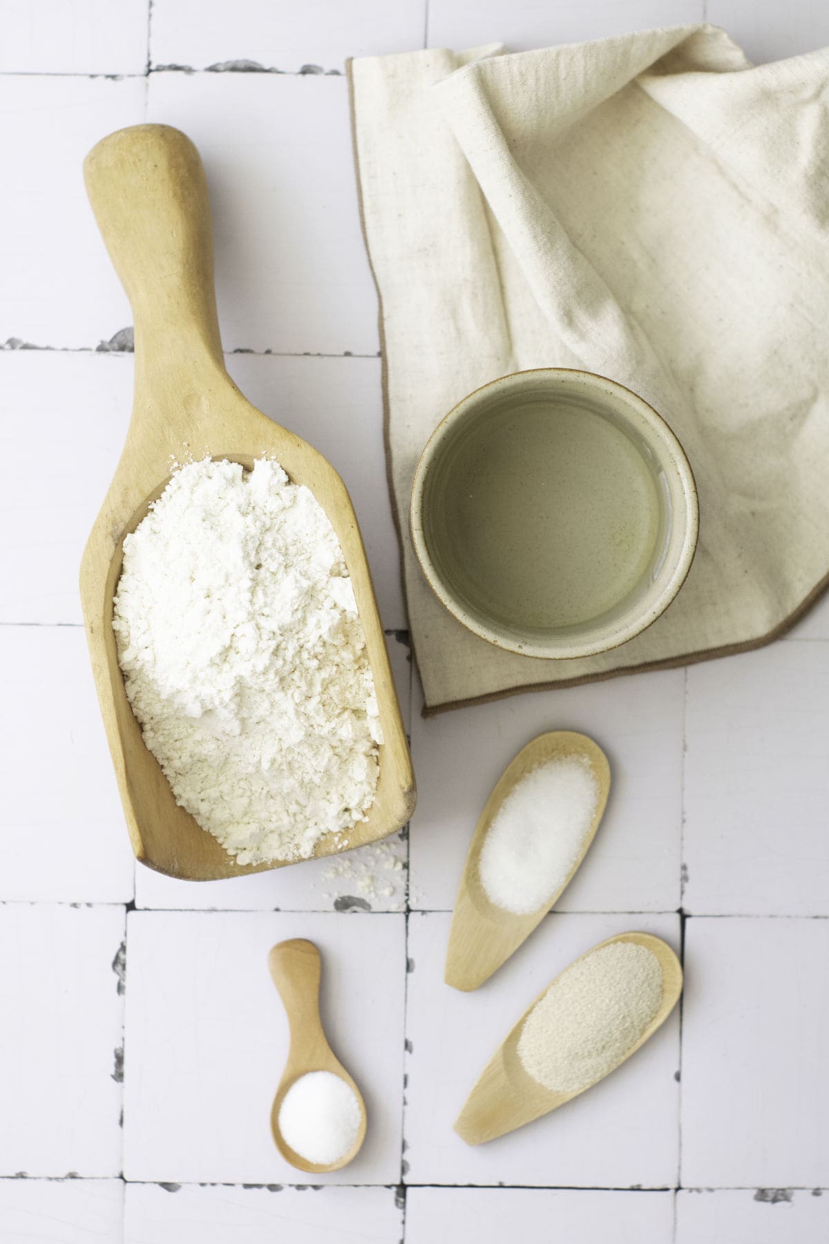 Wooden scoops with flour, sugar, and salt rest on a white tiled surface, reminiscent of ingredients for classic French baguettes. Nearby, a ceramic bowl and folded cloth complete the flat lay arrangement.