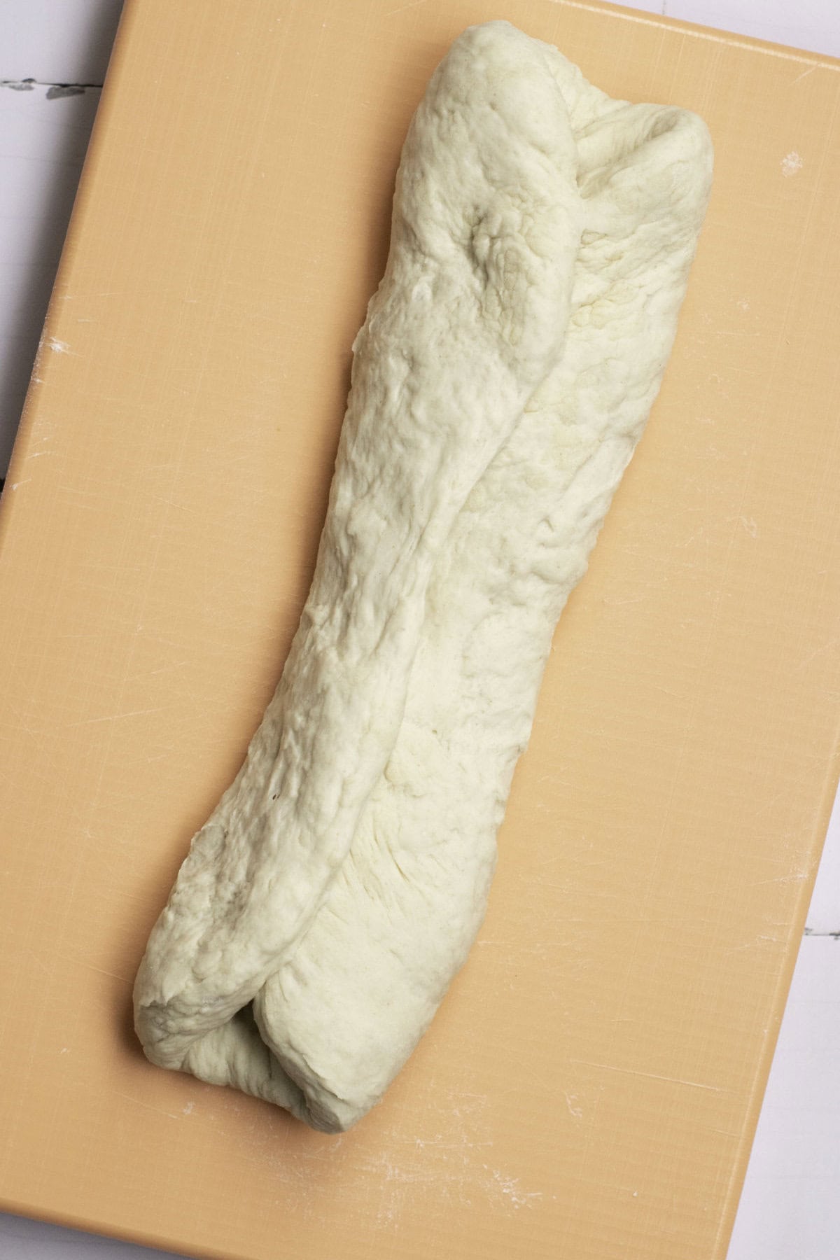 A long piece of bread dough, reminiscent of classic French baguettes, is twisted and placed on a wooden surface. The dough's light color and textured surface signal it's ready to be further shaped or baked.