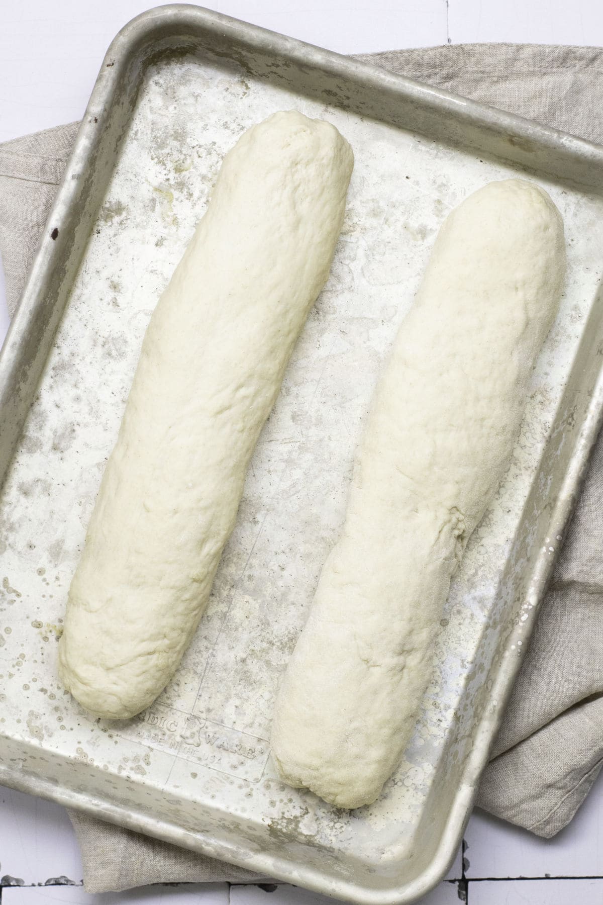 Two unbaked classic French baguettes rest on a metal baking sheet atop a light-colored cloth. The surface is lightly dusted with flour, and the dough is shaped into long, oval forms, ready for baking.