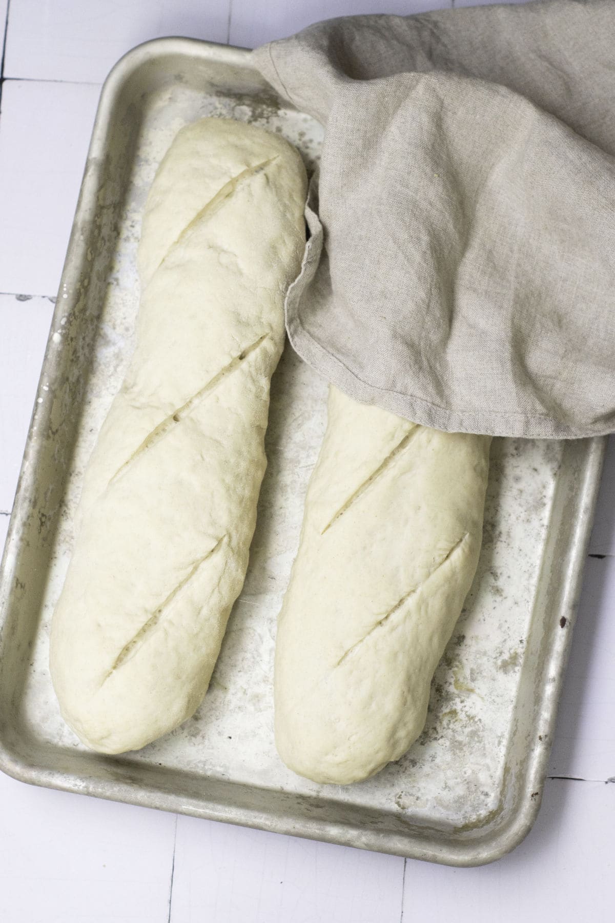 Two unbaked classic French baguette loaves with diagonal slits on top rest on a metal baking tray. A beige cloth partially covers one loaf, and the background is a white tiled surface.