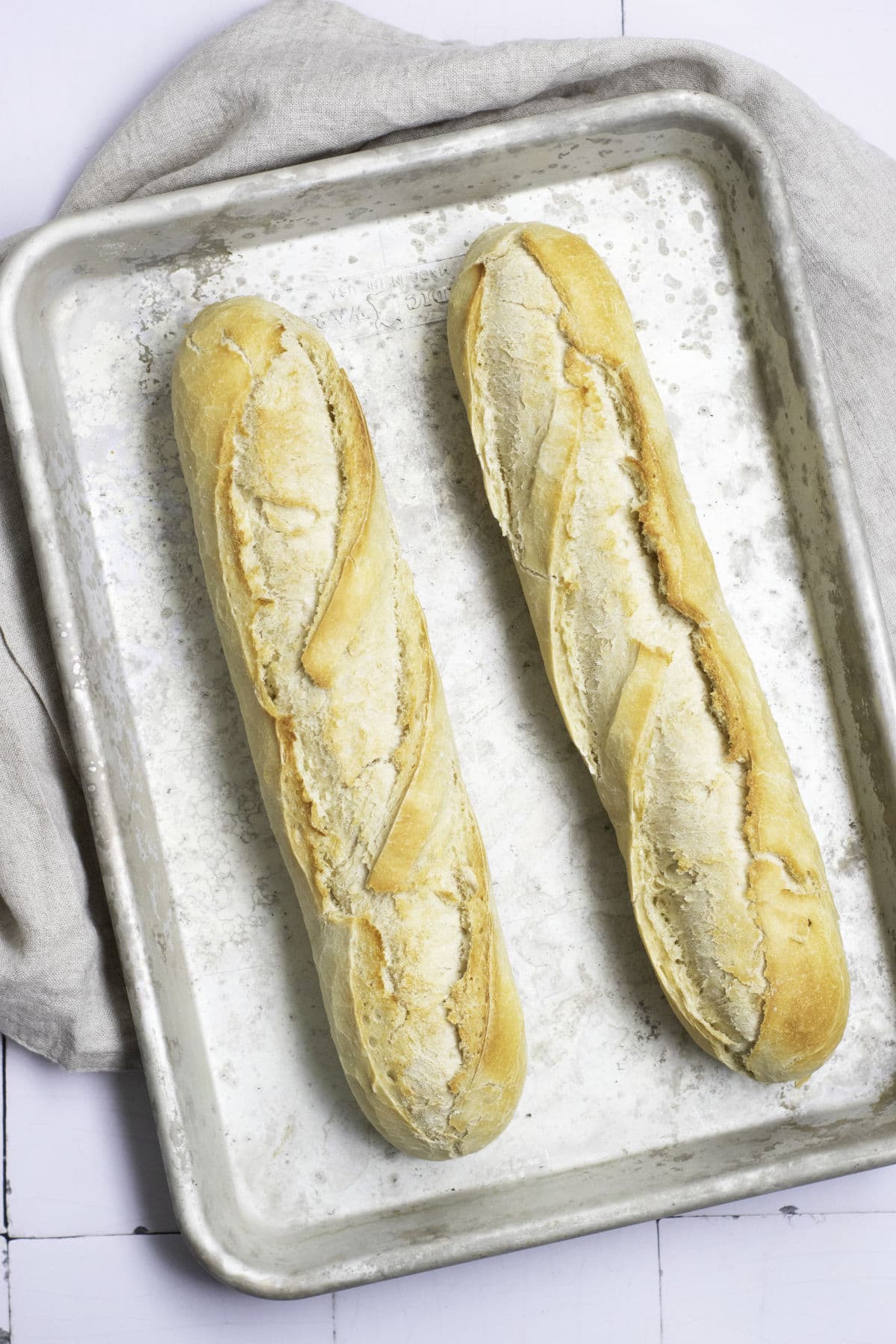 Two freshly baked classic French baguettes rest on a metal baking tray atop a light-colored cloth. Their golden crust boasts textured slashes, while beneath the tray lies a white, rustic wooden table.