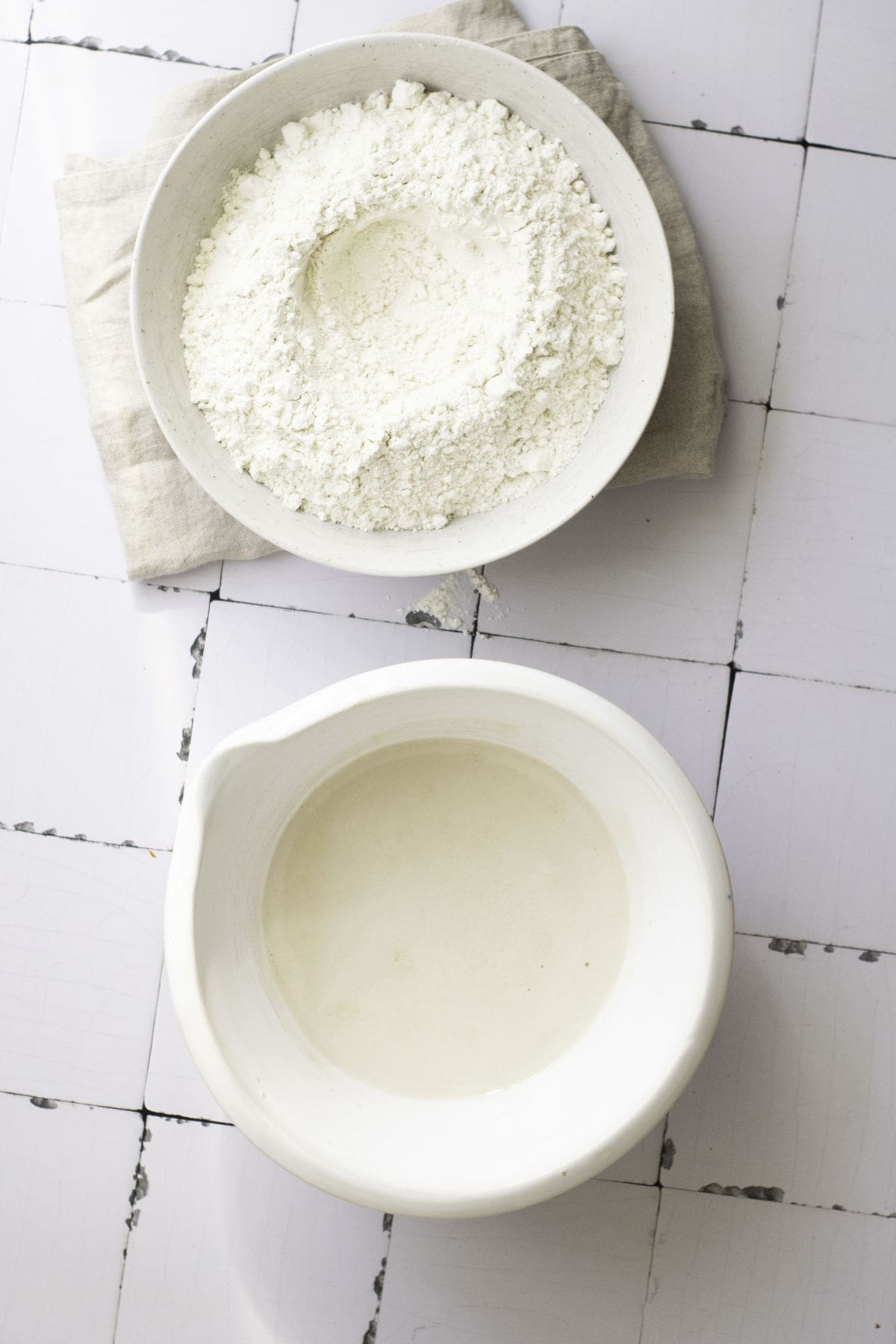 Two white bowls rest on a tiled surface; the top bowl cradles a mound of flour, essential for classic French baguettes, while the bottom bowl holds clear liquid. A folded cloth is partially visible beneath the top bowl.