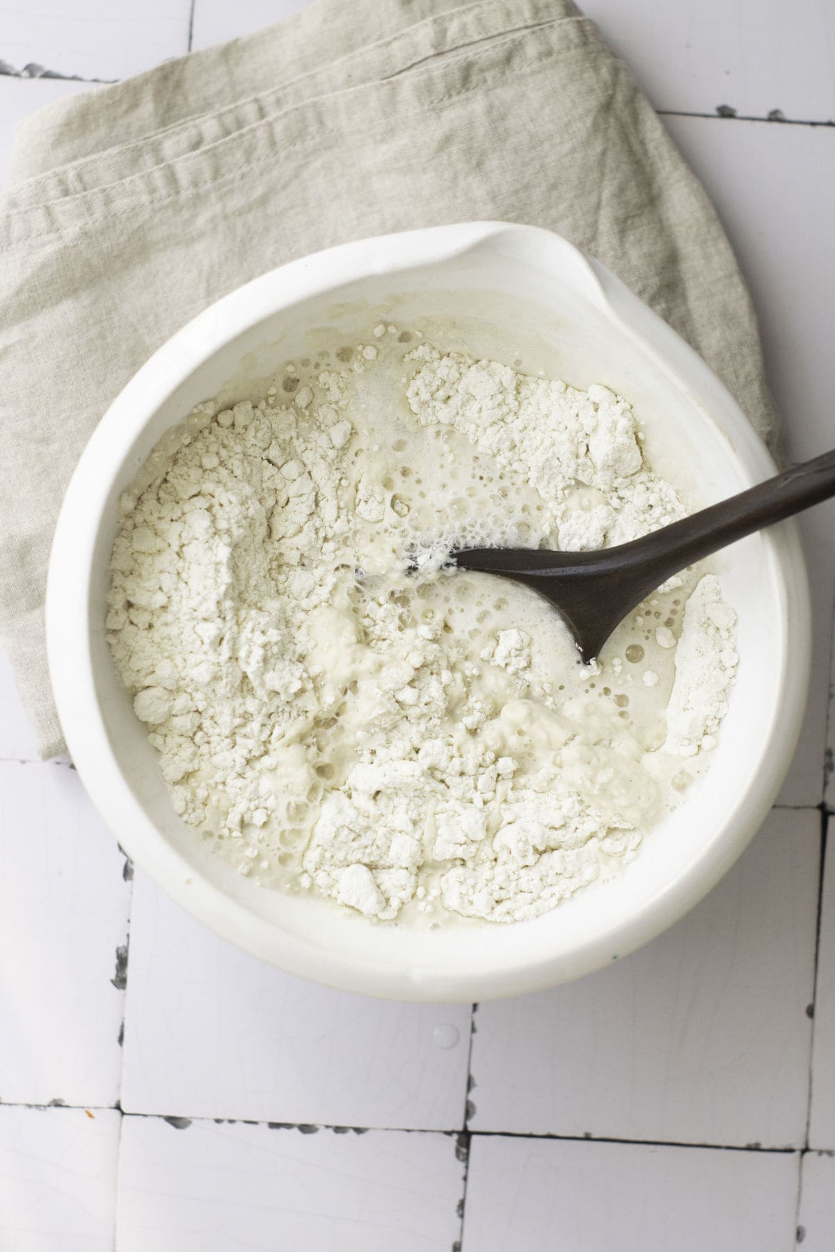 A white bowl filled with flour and water sits on a tiled surface, hinting at the beginning of classic French baguettes. A wooden spoon stands in the bowl, which rests on a light-colored cloth. The mixture appears to be the early stages of dough preparation.