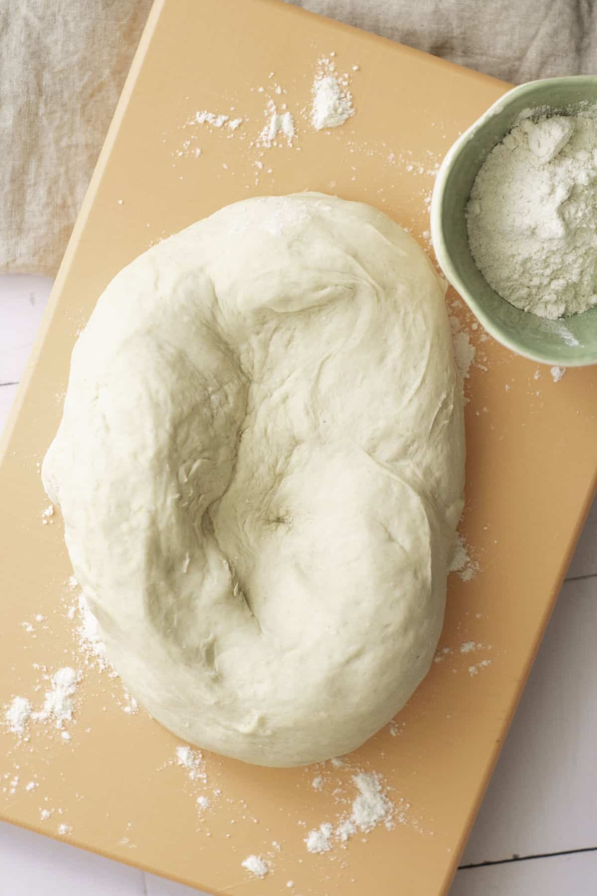 A ball of dough, destined to become classic French baguettes, rests on a light wooden cutting board, lightly dusted with flour. A small green bowl with flour awaits beside the dough. In the background, a textured beige cloth lies elegantly on a pristine white surface.