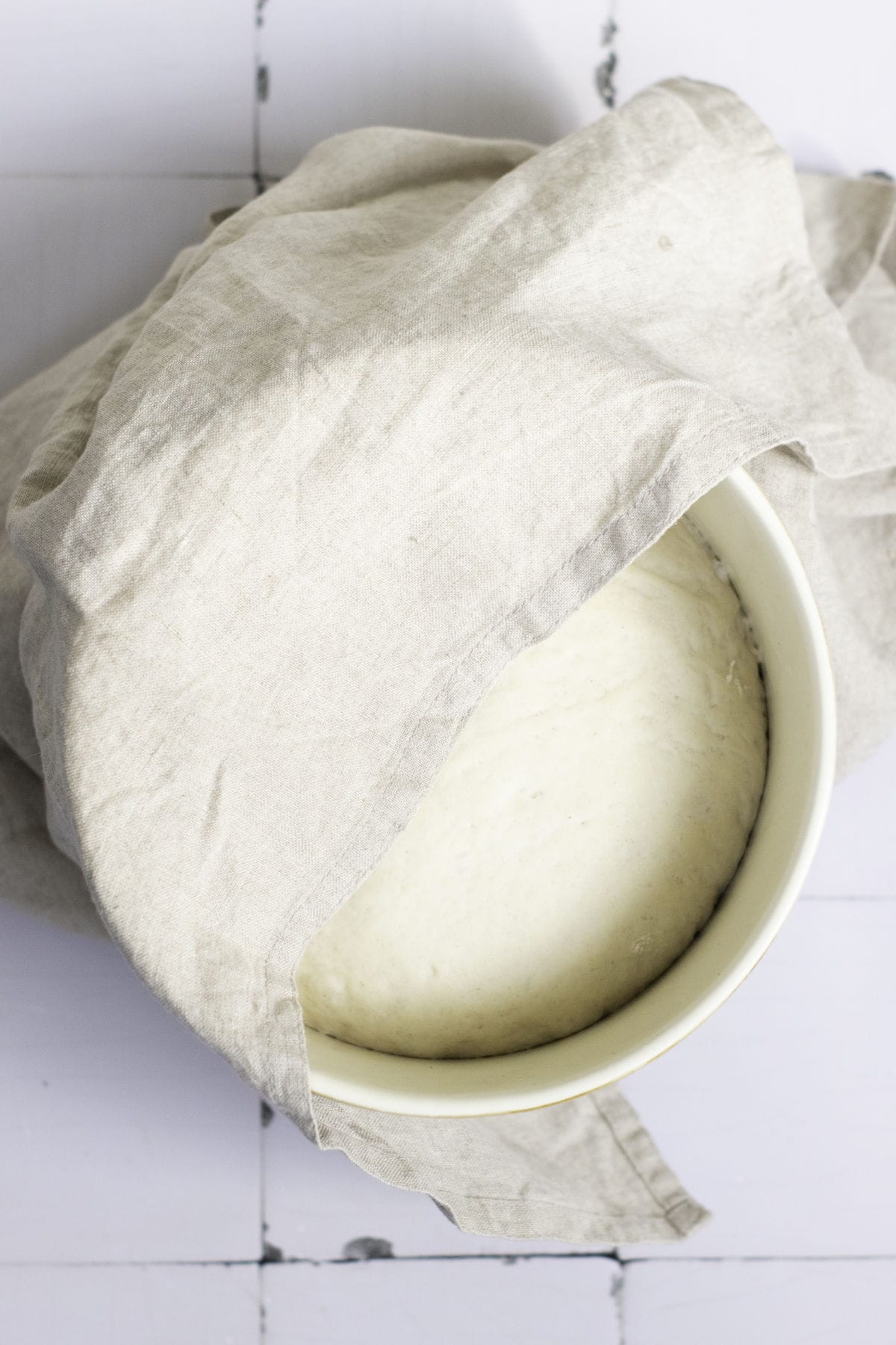 A bowl, covered with a beige cloth, cradles the rising bread dough destined to become classic French baguettes. Resting on a white surface with visible grid lines, the cloth gently veils part of its promising contents.