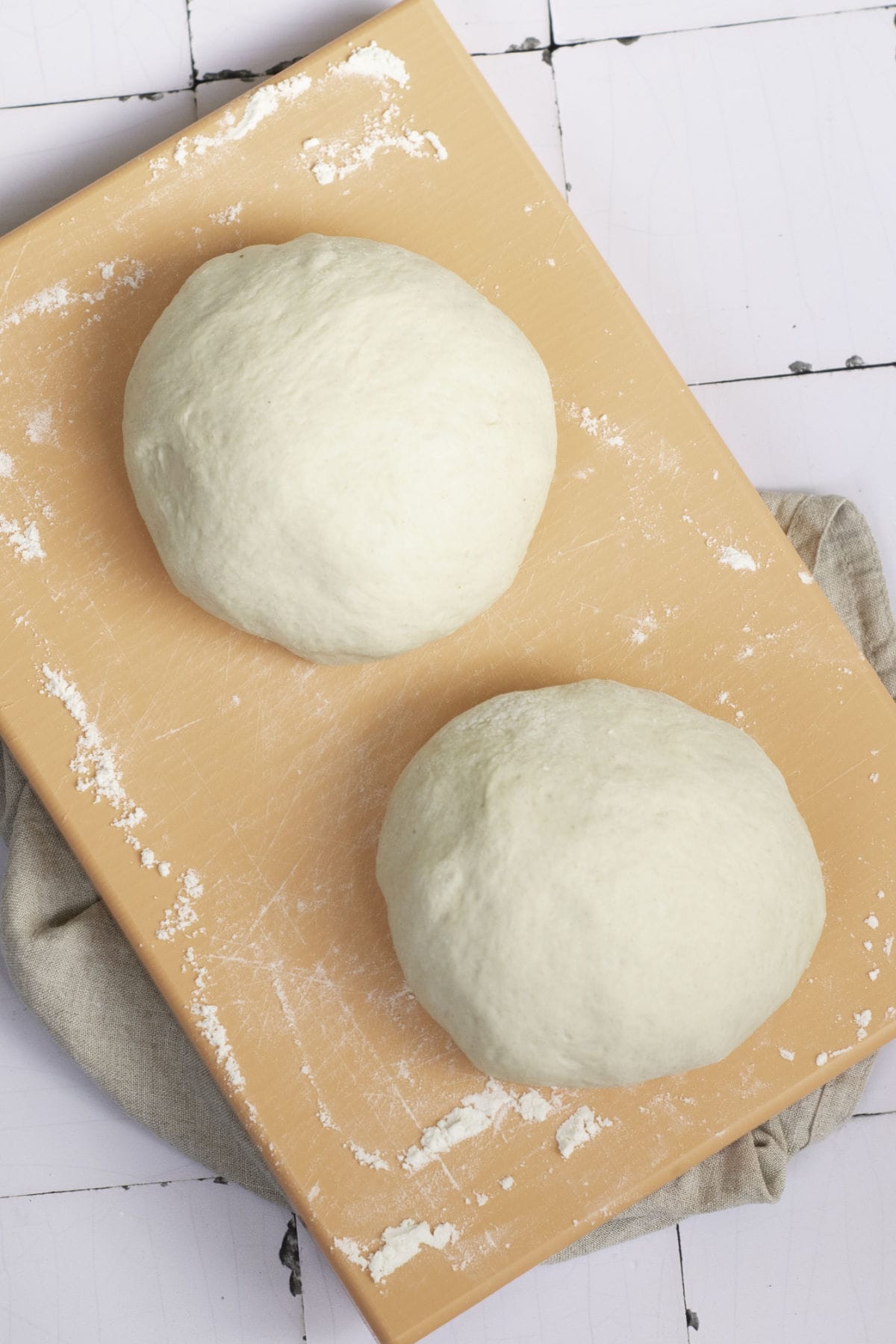 Two balls of pizza dough rest on a floured wooden board like classic French baguettes awaiting the oven. The board is placed on a light-colored cloth and a tiled surface. The dough is round and smooth, ready for preparation.