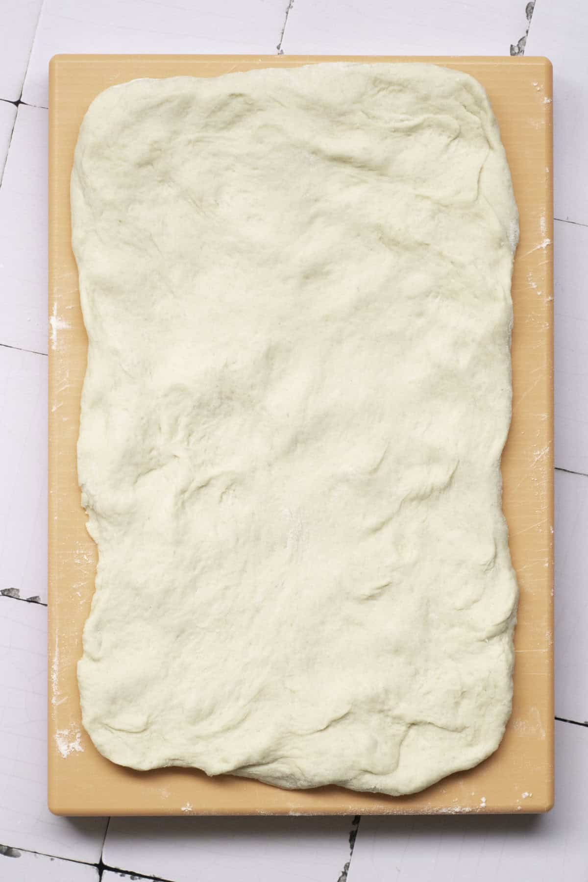 Rectangular pizza dough, reminiscent of classic French baguettes, is rolled out on a wooden board and placed on a white tiled surface.