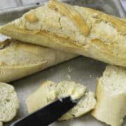 Classic French baguettes sit in the background as a sliced piece with butter spread on it takes center stage, a knife resting elegantly on top, all presented on a sleek metal tray.