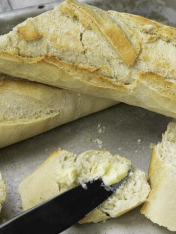 Classic French baguettes sit in the background as a sliced piece with butter spread on it takes center stage, a knife resting elegantly on top, all presented on a sleek metal tray.