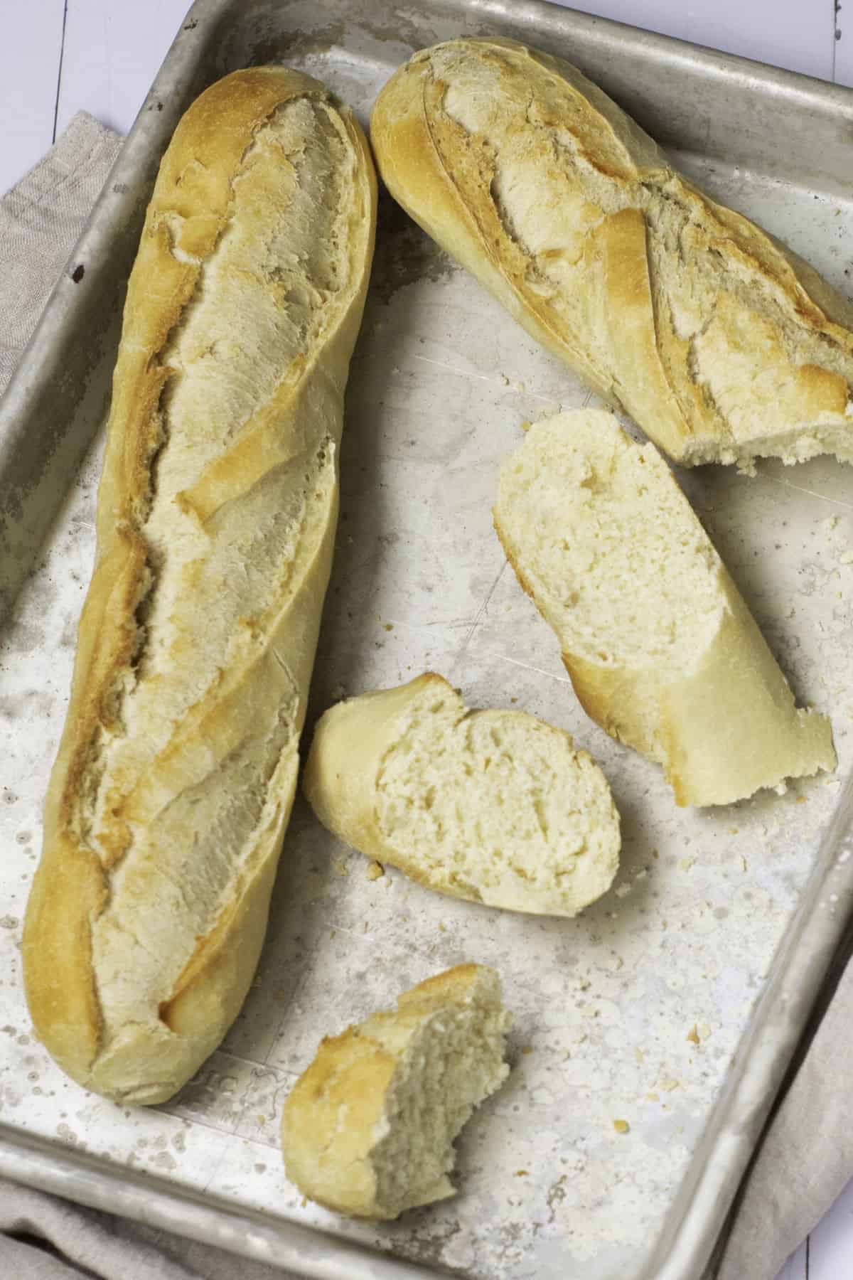 Two classic French baguettes and several cut pieces lie on a metallic baking tray. The baguettes are golden-brown with crispy crusts, and the texture inside appears soft and fluffy. A light-colored cloth is partially visible beneath the tray.