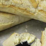 Close-up of freshly baked classic French baguettes with a cracked, golden crust. One baguette is sliced open with butter being spread on it using a knife. The background is a metal surface.
