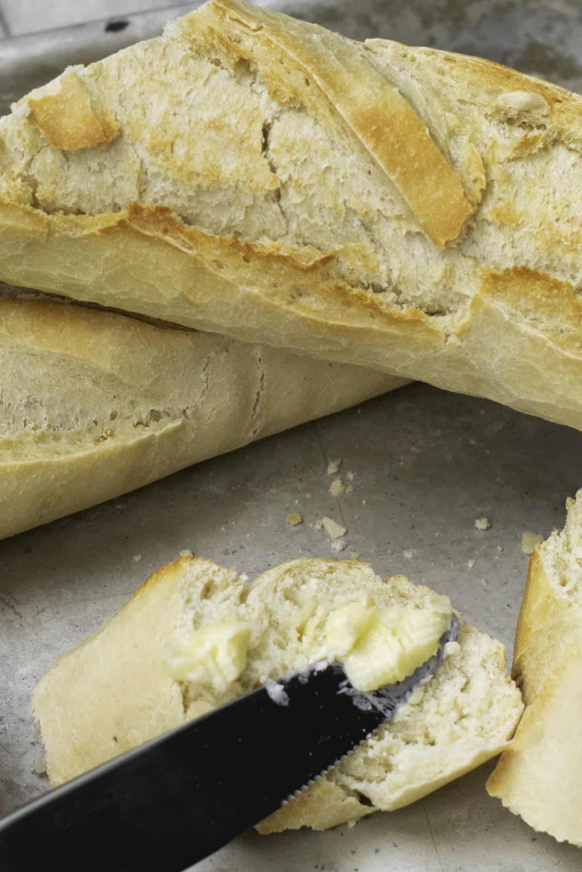 Close-up of freshly baked classic French baguettes with a cracked, golden crust. One baguette is sliced open with butter being spread on it using a knife. The background is a metal surface.