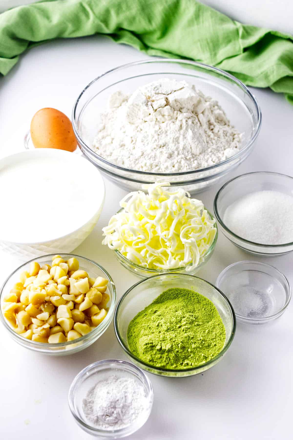 An assortment of baking ingredients in glass bowls, ideal for making matcha green tea scones—flour, sugar, grated butter, chopped chestnuts, matcha powder, baking powder, salt, a bowl of liquid, and an egg—arranged on a white surface with a green cloth.