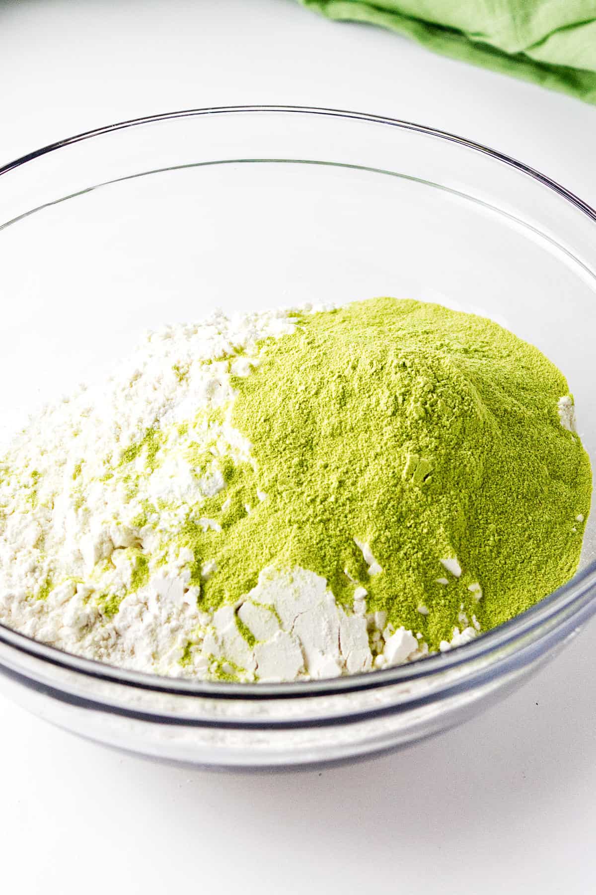A clear glass bowl contains a mixture of white flour and bright green matcha powder, ready to be blended for delicious matcha green tea scones, on a white surface with a green cloth in the background.