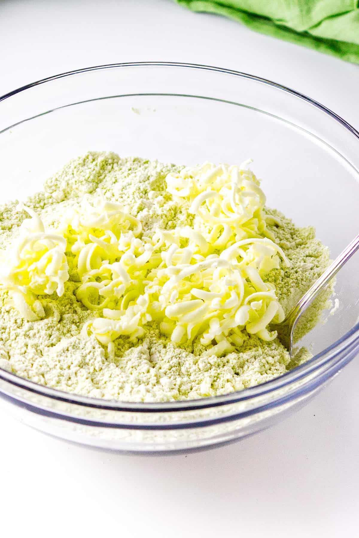 A glass bowl filled with green-tinted flour mixture and shredded butter for matcha green tea scones, with a metal spoon on the side, on a white surface.