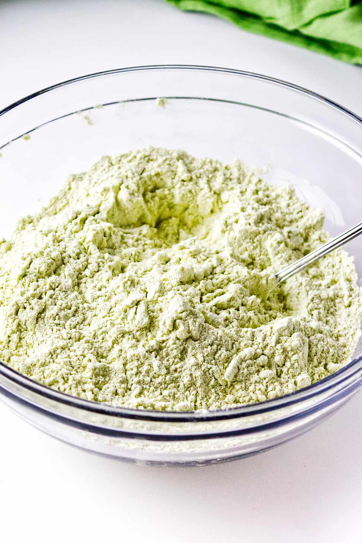 A clear glass bowl filled with a pale green powdery mixture for matcha green tea scones sits on a white surface, with a metal spoon partially buried in the mixture. A green cloth is visible in the background.