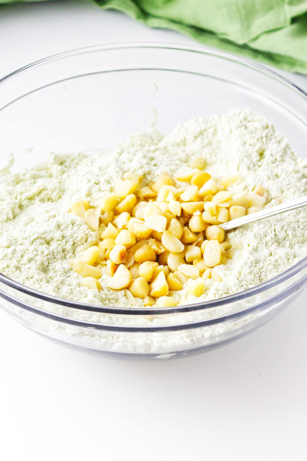 A clear glass bowl containing flour mixture with white chocolate chips on top, being stirred with a metal spoon—perfect for making matcha green tea scones. A green cloth is partly visible in the background.