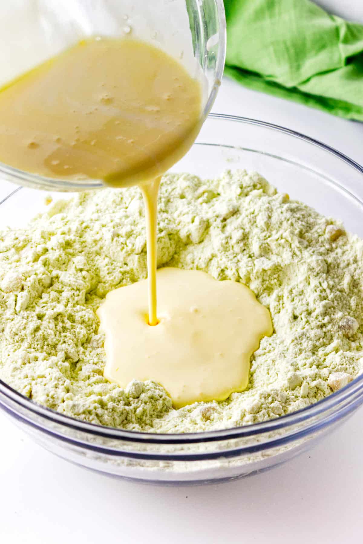 A glass bowl filled with a greenish flour mixture for matcha green tea scones has a well in the center, while yellow liquid is being poured in from a measuring cup. A green cloth appears in the background.