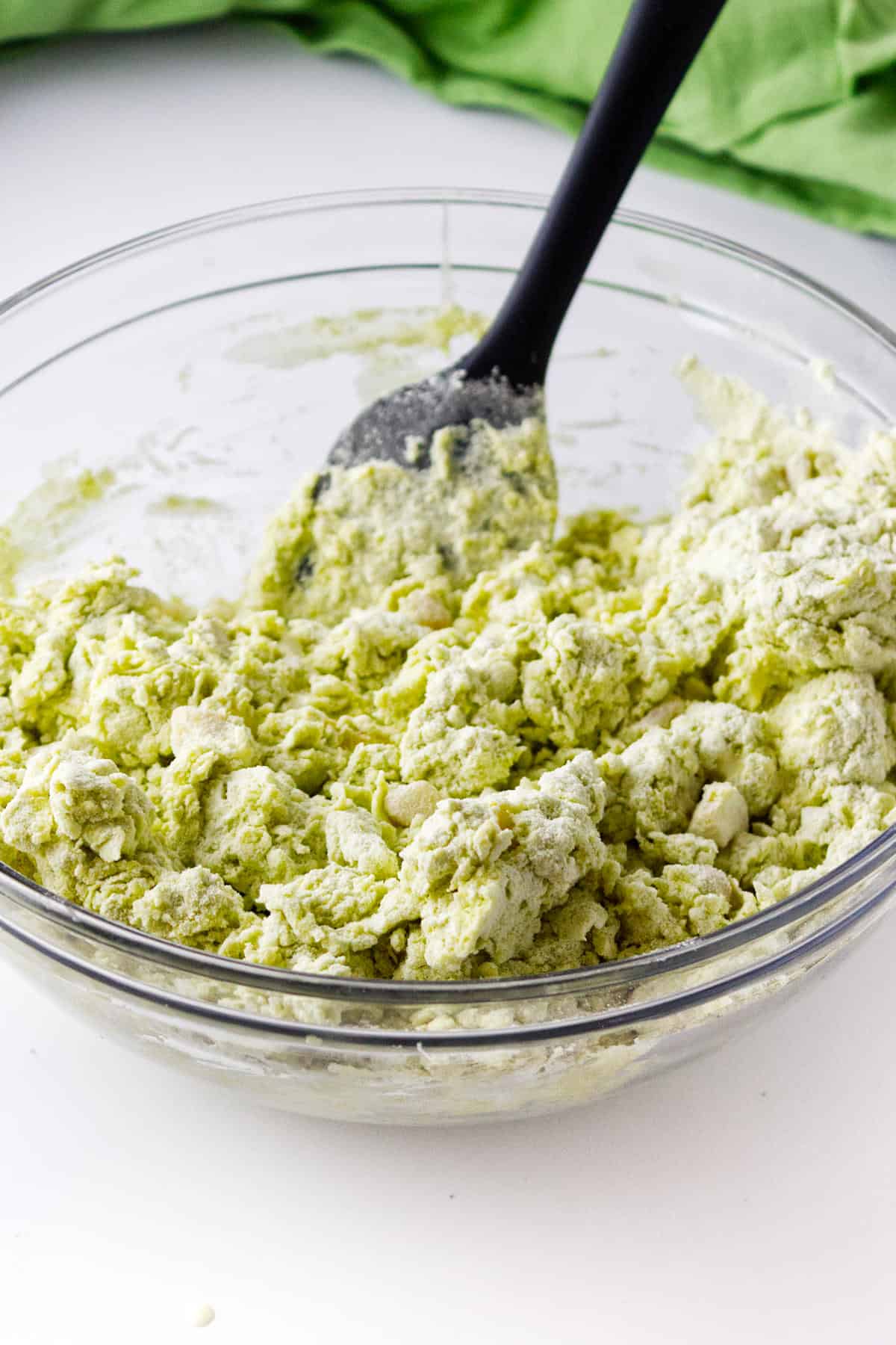 A clear glass bowl filled with chunky, pale green matcha green tea scone dough is being stirred with a black spatula on a white surface. A green cloth is visible in the background.
