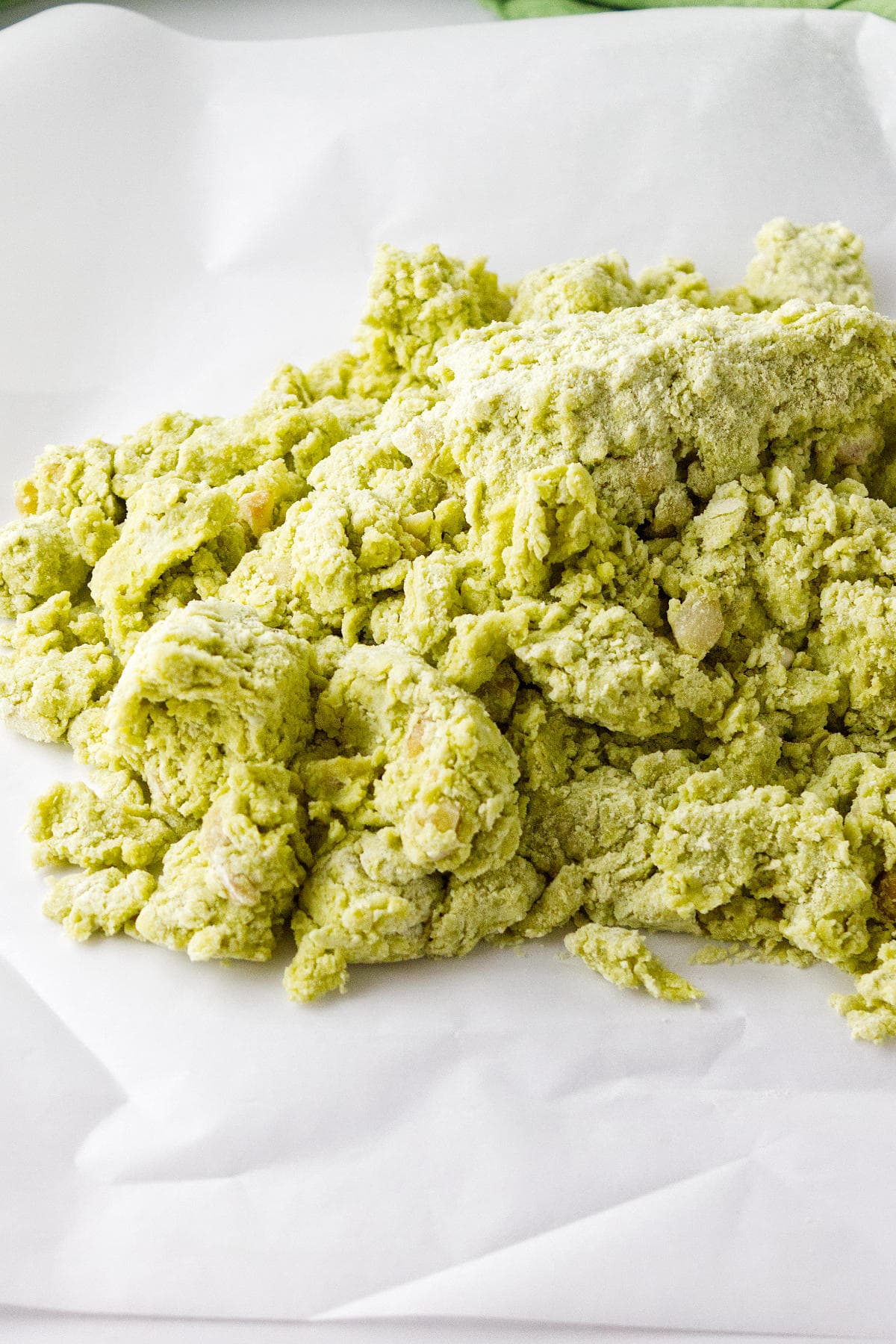 A close-up of a mound of green, crumbly dough for matcha green tea scones on a sheet of white parchment paper.