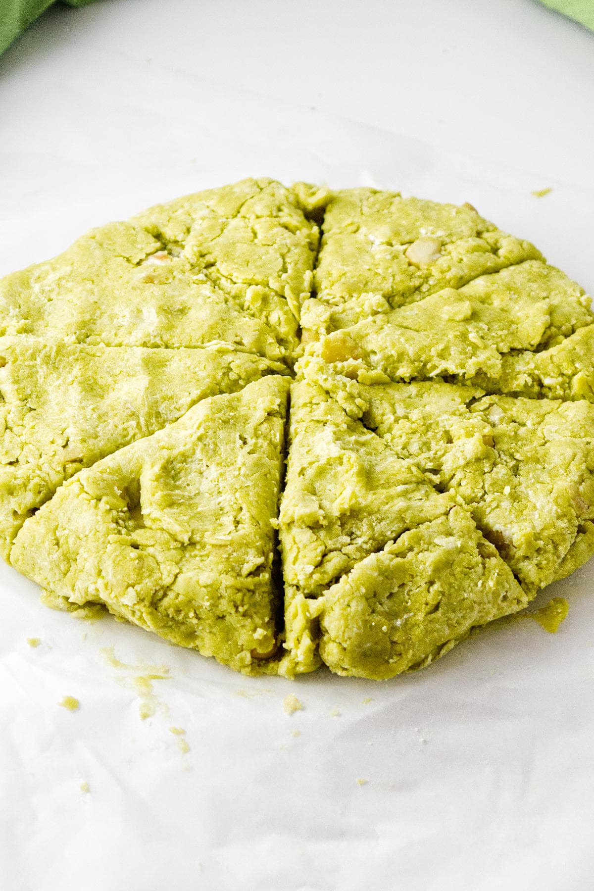 A round, greenish dough for matcha green tea scones has been cut into six triangular wedges, resting on white parchment paper. The dough appears crumbly and textured, with some small chunks visible.