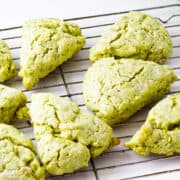 Eight matcha green tea scones rest on a metal cooling rack, their cracked surfaces and triangular shapes highlighted against a clean white background.