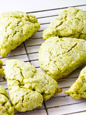 Eight matcha green tea scones rest on a metal cooling rack, their cracked surfaces and triangular shapes highlighted against a clean white background.
