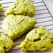 Four matcha green tea scones sit on a metal cooling rack with a white background and a green cloth in the corner. The scones have a slightly rough, crumbly texture.