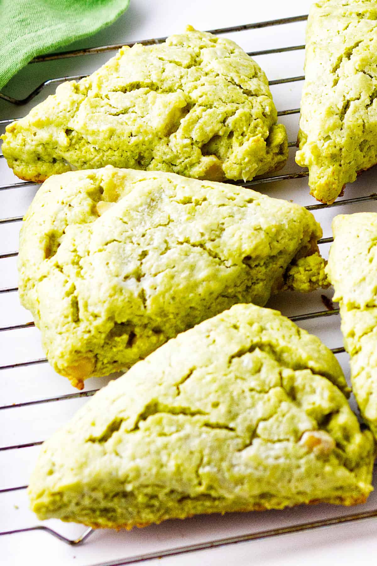 Four golden-green matcha green tea scones rest on a wire cooling rack, with one partially overlapping another. The scones have a crumbly texture, and a green cloth is visible in the background.
