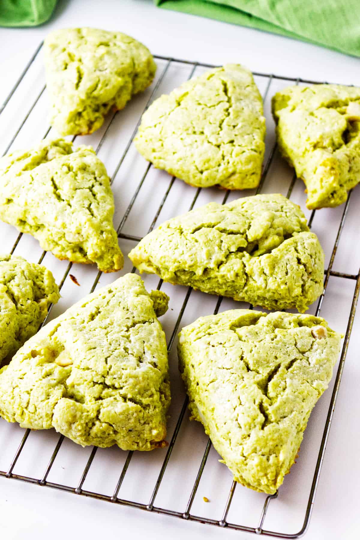 Eight matcha green tea scones are arranged on a wire cooling rack, with a green cloth visible in the background. The triangular scones have a slightly crumbly texture.