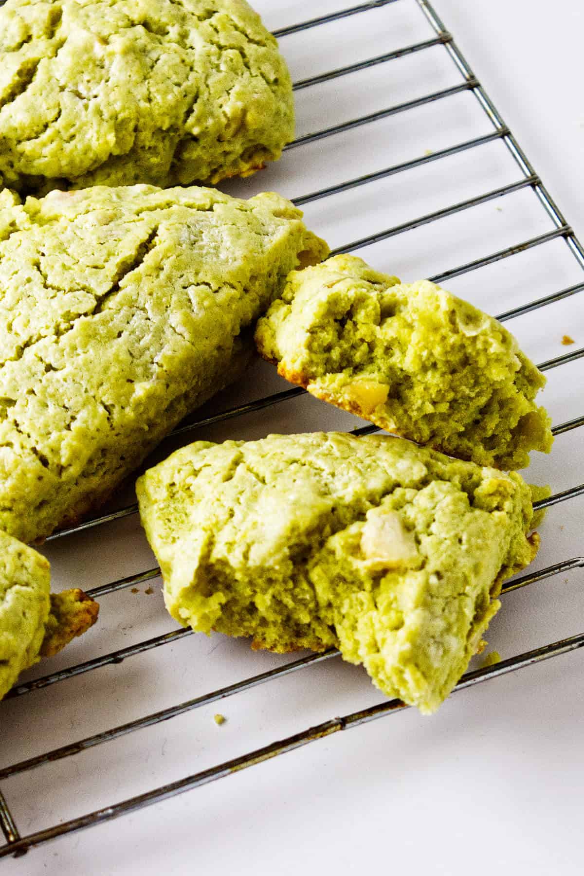 Three matcha green tea scones rest on a metal cooling rack, with one scone broken in half to reveal its moist, crumbly interior. The background is a clean, white surface.