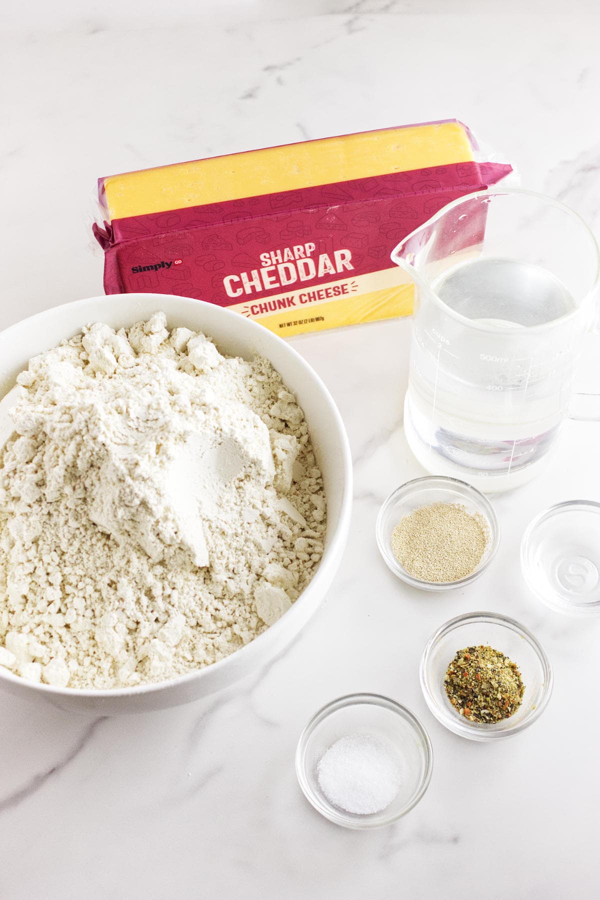 A bowl of flour is surrounded by ingredients for baking no-knead cheese bread, including a block of sharp cheddar cheese, a measuring cup of water, yeast, salt, and a small dish of herbs on the white marble surface.