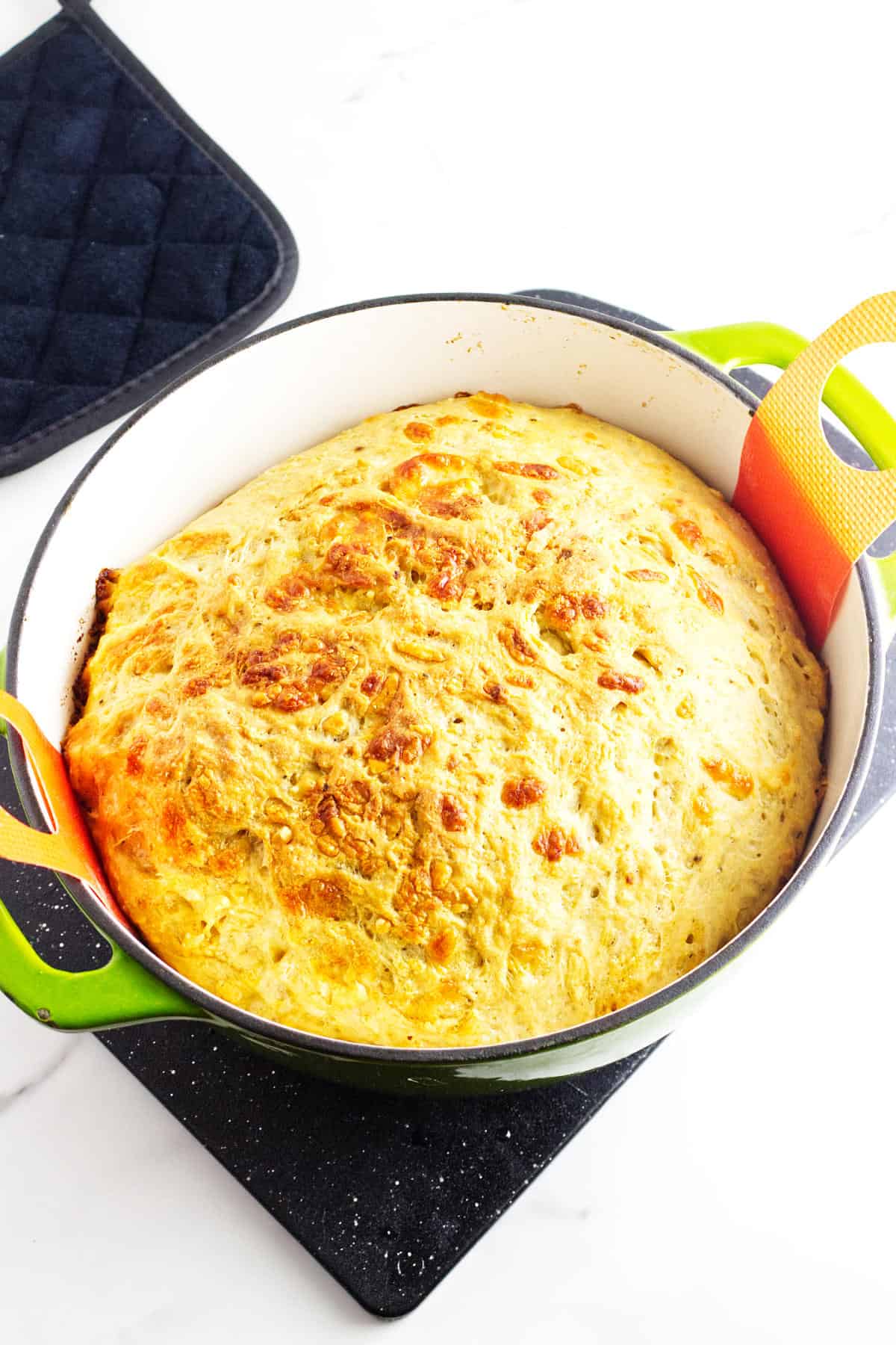 A freshly baked no knead cheese bread loaf in a green Dutch oven rests on a black trivet. The bread has a golden, crispy crust, infused with cheesy goodness, and there is a navy blue oven mitt nearby. The scene is set on a white surface.