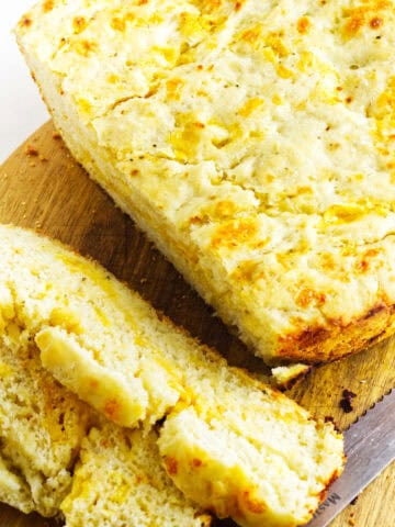 A loaf of no-knead cheese bread rests on a wooden cutting board, partially sliced. The bread features a rough texture with a golden, cheesy crust and visible cheese bits throughout. A knife is placed beside the board on a white surface.