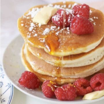A stack of fluffy pancakes topped with butter, fresh raspberries, powdered sugar, and syrup, served on a white plate with extra raspberries on the side.