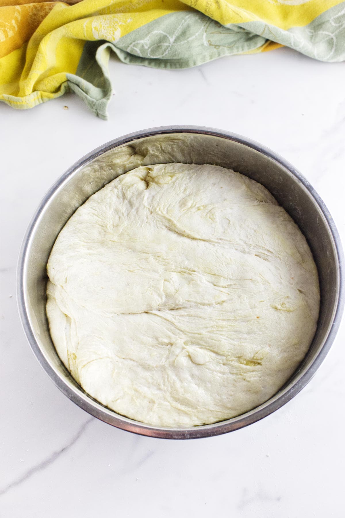 A metal bowl filled with risen white sourdough discard sandwich bread dough sits on a white countertop, with a yellow and green kitchen towel partially visible in the background.