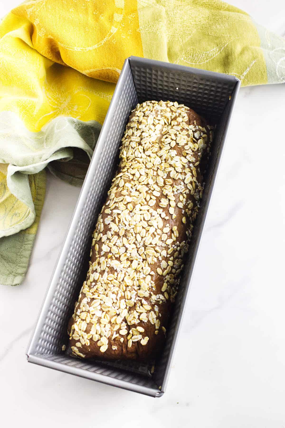 A loaf of sweet pumpernickle bread topped with oats sits in a rectangular metal baking pan, with a yellow and green kitchen towel nearby on a white surface.