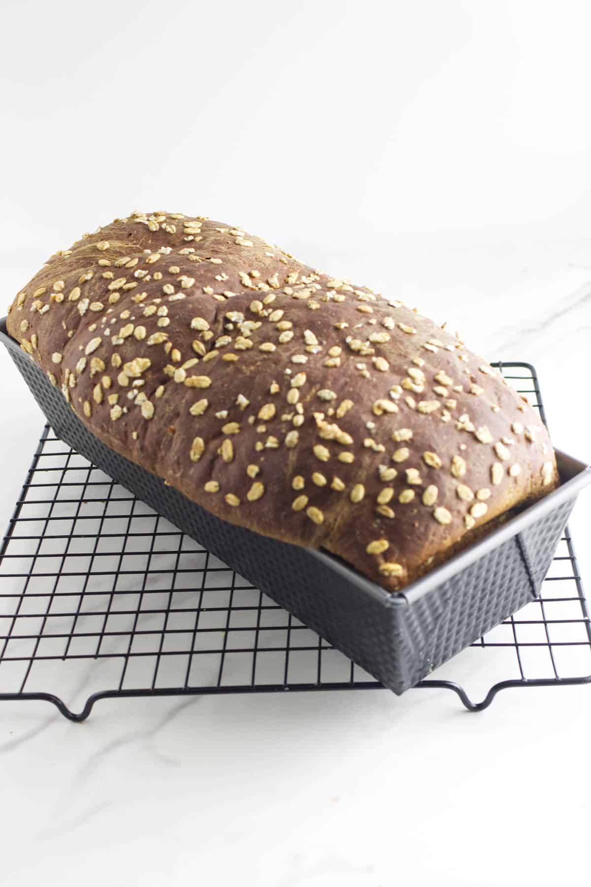 A loaf of Cheesecake Factory brown bread, oat-topped and sweet like pumpernickle bread, sits in a baking pan on a black wire cooling rack against a white background.