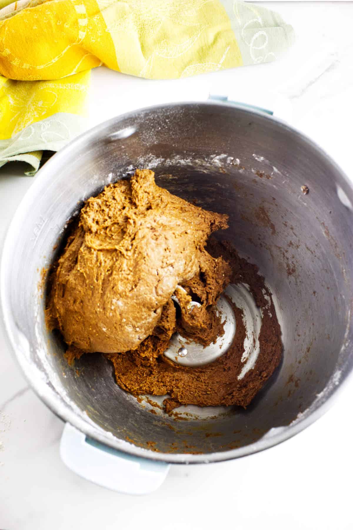 A large metal mixing bowl contains thick, brown dough for sweet pumpernickle bread. Part of the dough is stuck to the side of the bowl. In the background, a yellow and green kitchen towel rests on a white surface.