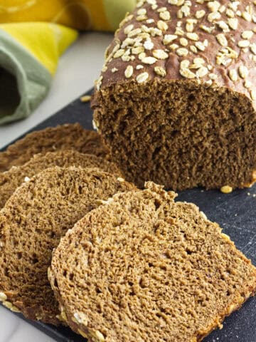 A loaf of Cheesecake Factory brown bread sits on a black cutting board, with oat topping and several slices cut and arranged in front. A yellow and green cloth is partially visible in the background.