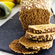 A loaf of dark brown sweet pumpernickle bread sits on a black cutting board, with several slices fanned out in front. The bread, reminiscent of Cheesecake Factory brown bread, is topped with rolled oats; a cloth napkin rests in the background.
