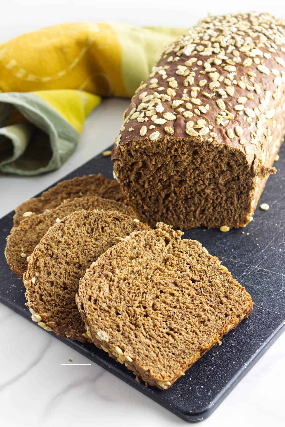 A loaf of sweet pumpernickle bread topped with oats sits on a black cutting board. Several slices are cut and laid flat in front of the loaf. A yellow and green cloth is partially visible in the background.